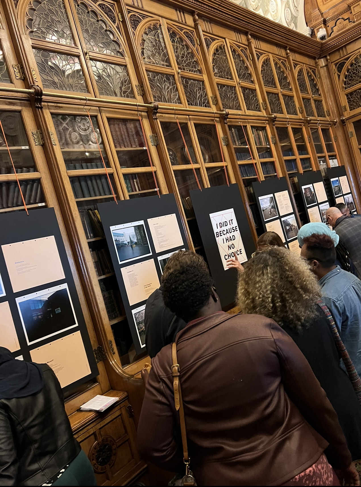 People viewing an exhibition in a room with images and text mounted on boards propped up against book cabinets